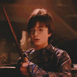 May include: A photographic print featuring a young person with dark hair and round glasses, wearing a plaid shirt. The individual is holding a thin wooden wand in their right hand. A dark, handwritten signature is overlaid across the center of the image.