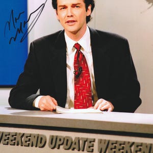 May include: A glossy photograph featuring a news anchor wearing a black suit, white collared shirt, and a red patterned tie. The anchor sits behind a desk labeled WEEKEND UPDATE WEEKEND. A black ink signature is visible on the left side of the image.