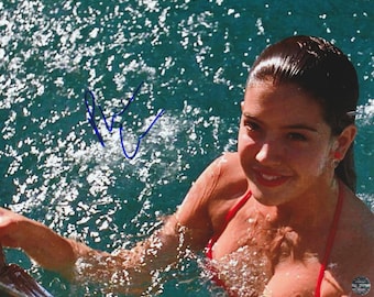 May include: A glossy photograph featuring a person with dark hair in a red bikini top, partially submerged in sparkling blue water. A blue ink signature is visible on the water surface to the left of the person, and a small circular logo is in the corner.