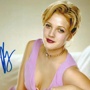 May include: A portrait of a person with short blonde hair, wearing a light purple sleeveless top and a gold-toned choker necklace. The person has dark pink lipstick and is looking toward the camera. A blue ink signature is visible on the left side.