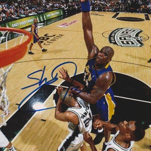 May include: A basketball player in a purple and gold jersey shoots the ball during a game. Two other players in black and white jerseys attempt to block the shot. The court has the words "San Antonio Spurs" and "BC Center" on it.