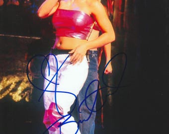 May include: A photograph of a woman on stage holding a microphone. She wears a shiny pink tube top and white pants with pink accents. Her blonde hair is pulled back, and she is singing. A person is partially visible behind her.