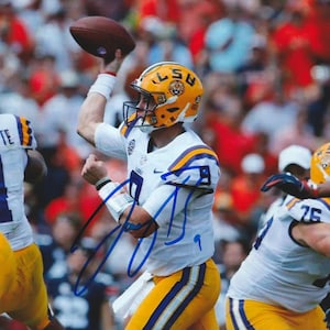 May include: A football player in a white, yellow, and purple uniform throws a brown football. The player wears a yellow helmet with the letters "LS" and a purple and yellow logo. The background is blurred with other players and spectators.