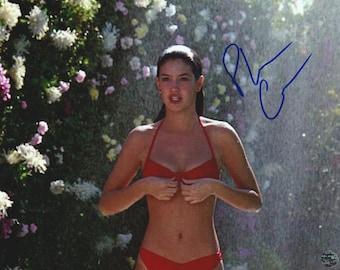 May include: A glossy photograph featuring a person wearing a red bikini top and bottom, standing in front of a wall of white and pink flowers. The image includes a blue ink signature on the right side and a small circular authentication seal.