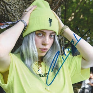 May include: A person wearing a neon green beanie with a black silhouette of a person on it. They are also wearing a yellow t-shirt and gold jewelry. The person has silver hair and is holding their head with both hands.