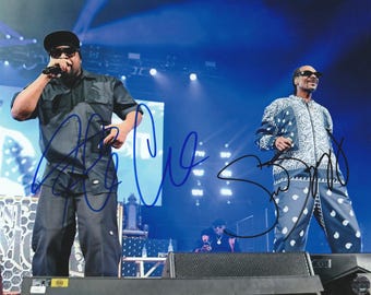 May include: A concert photograph featuring Ice Cube in a black short-sleeved shirt and pants, and Snoop Dogg in a blue and white paisley patterned zip-up jacket and matching pants. Both performers wear sunglasses, with blue ink autographs across the image.