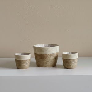 Może przedstawiać: Trzy tkane donice o różnych rozmiarach. Donice mają naturalną brązową podstawę i kremową górę. Każda donica ma plastikową wyściółkę w środku. Donice są ustawione na białej powierzchni.