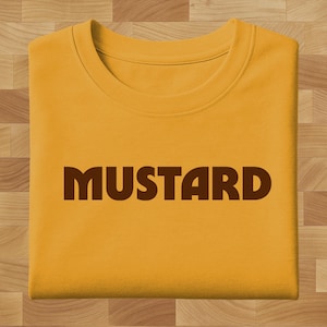 Puede incluir: Sudadera de color mostaza con la palabra "MUSTARD" impresa en letras oscuras de bloque marrón. La camiseta de cuello redondo está doblada cuidadosamente sobre una superficie de madera.