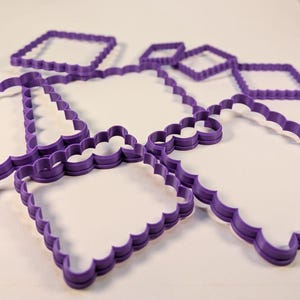 May include: A collection of purple plastic cookie cutters in various shapes, including squares and rectangles, all featuring a scalloped edge design. The cutters are arranged in a cluster, showcasing their distinct geometric outlines and uniform purple color.