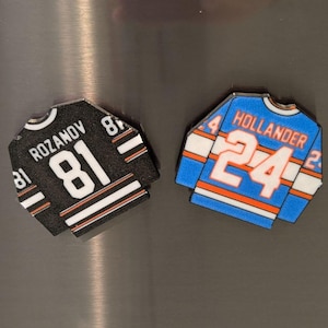 May include: Two hockey jersey-shaped magnets. One is black with white and orange stripes, the name "ROZANOV" and the number "81". The other is blue with orange and white stripes, the name "HOLLANDER" and the number "24".