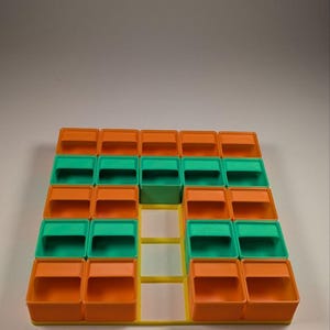 May include: A colorful storage organizer with a grid of small, square compartments. The compartments are orange, green, and yellow, arranged in a geometric pattern. The organizer is made of plastic and is designed for small items.