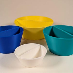 May include: Four colorful, divided bowls in blue, yellow, teal, and white. Each bowl has a single divider creating two compartments. The bowls are made of a smooth material and are arranged on a light background.