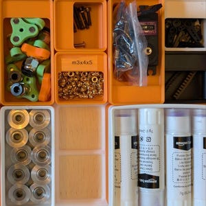 May include: A collection of small parts and supplies organized in orange and white plastic containers. Items include screws, nuts, gears, and glue sticks. Some containers have labels, including "m3x4x5".
