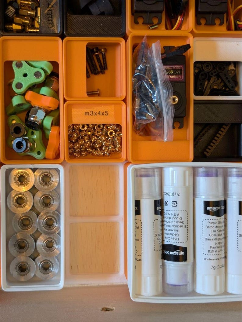 May include: A collection of small parts and supplies organized in orange and white plastic containers. Items include screws, nuts, gears, and glue sticks. Some containers have labels, including "m3x4x5".