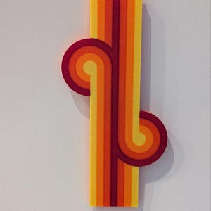 May include: A retro-style decorative piece featuring a vertical yellow and orange striped rectangle with two red and orange swirl designs. The piece has a 1970s aesthetic and is set against a white background.