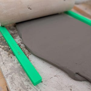 May include: A light brown wooden rolling pin rests on a flat slab of dark gray clay. Two bright green plastic guide sticks, labeled SECOND CRYSTAL STANDARD 2016, are positioned on either side of the clay to ensure an even thickness.