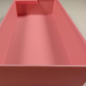 May include: A light pink rectangular container with a small raised section on one side. The interior has a textured, ridged pattern. A yellow measuring tape is visible in the background, showing a measurement of approximately 2 inches.
