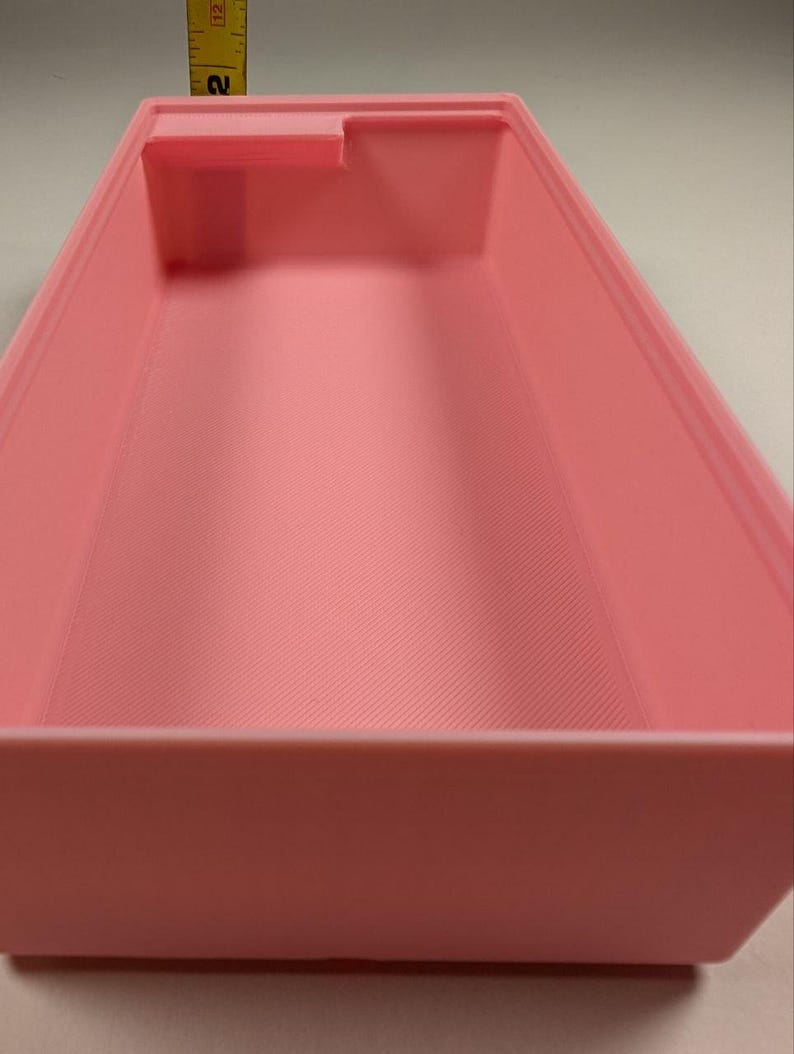 May include: A light pink rectangular container with a small raised section on one side. The interior has a textured, ridged pattern. A yellow measuring tape is visible in the background, showing a measurement of approximately 2 inches.