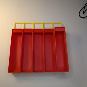 May include: A wall-mounted organizer with five red rectangular compartments and a yellow top bar. The compartments are open at the top and appear to be made of plastic. The organizer is mounted on a white wall.