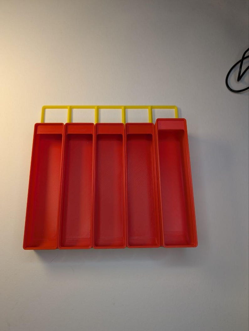 May include: A wall-mounted organizer with five red rectangular compartments and a yellow top bar. The compartments are open at the top and appear to be made of plastic. The organizer is mounted on a white wall.