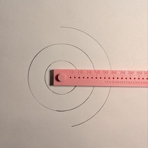 May include: A pink plastic measuring tool with a circular pivot point and a straight edge featuring numerical markings from 10 to 90 and the text "SECONDCITYSTANDARD" is positioned over three concentric black circles drawn on a white surface.