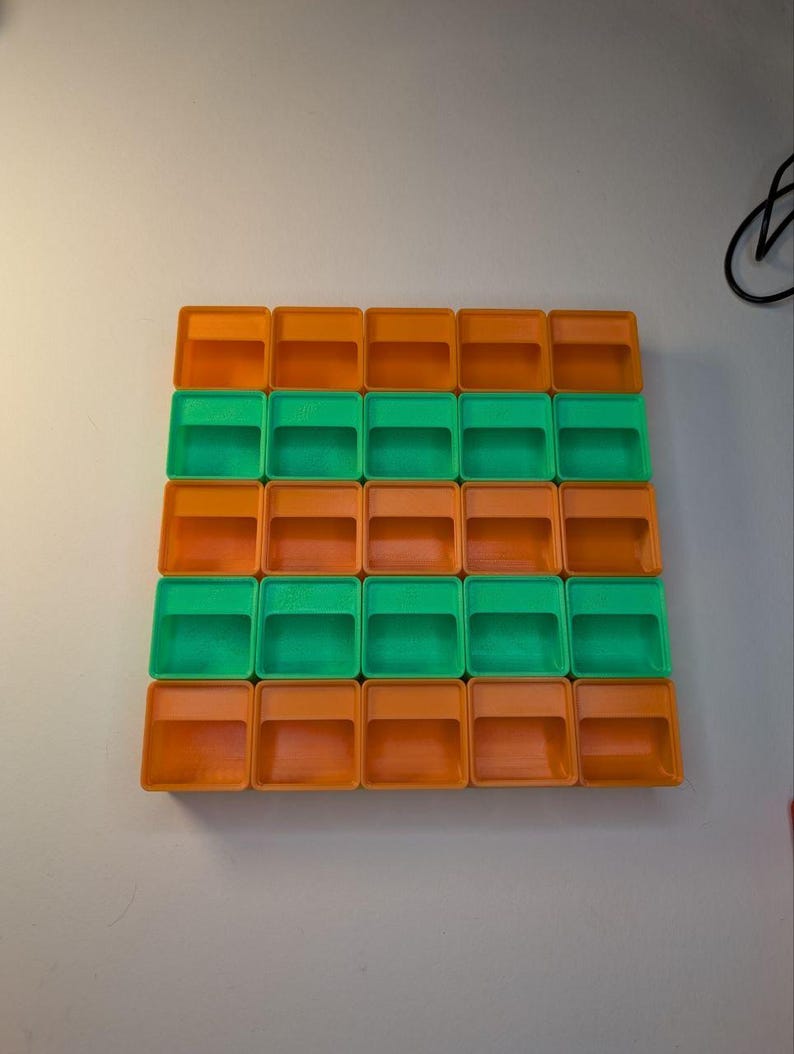 May include: A grid of twenty-four small, square, plastic containers. The containers alternate between orange and green, arranged in a pattern of four rows and six columns. The containers are empty and have a simple, open design.