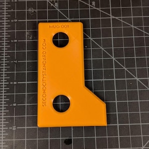 May include: Bright orange, L-shaped tool with two circular holes. The text "MUG 001" and "SECONDCITYSTANDARD.COM" are printed on the tool. The tool is on a black grid surface.