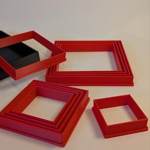 May include: A collection of red square-shaped cookie cutters in various sizes. The cutters are made of plastic and are arranged on a white surface. A black square container is visible in the background.