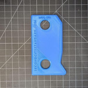 May include: A bright blue, rectangular tool with two circular holes and a unique shape. The text "MUG 010" and "SECONDCITYSTANDARD.COM" are printed on the surface. The tool is likely 3D-printed.
