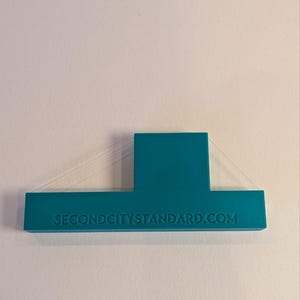 May include: A teal, T-shaped plastic object with a flat base and a rectangular vertical extension. The text SECONDCITYSTANDARD.COM is embossed in capital letters across the front of the base. Thin, translucent support filaments are visible.