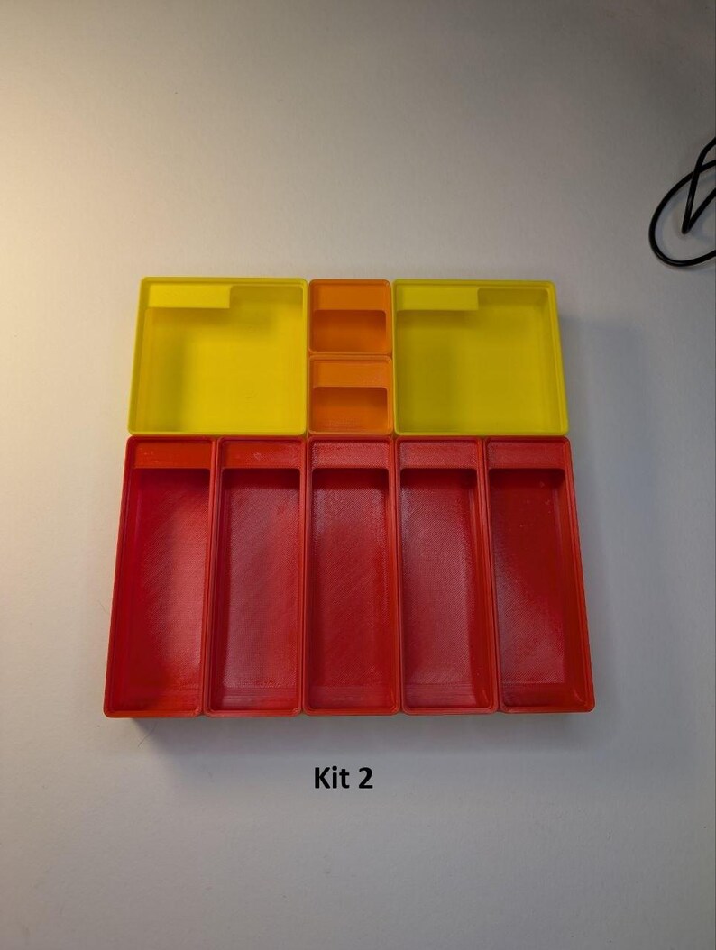 May include: A set of colorful plastic storage containers. The set includes yellow and orange square containers on top, and red rectangular containers below. The text "Kit 2" is printed at the bottom.