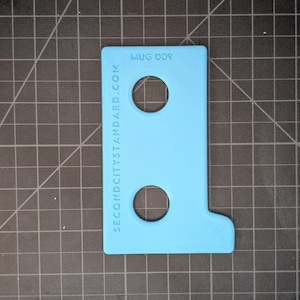 May include: A light blue plastic tool with two circular holes and a right-angle extension. The text "MUG 009" and "SECONDCITYSTANDARD.COM" are printed on the surface. The tool is on a black cutting mat.