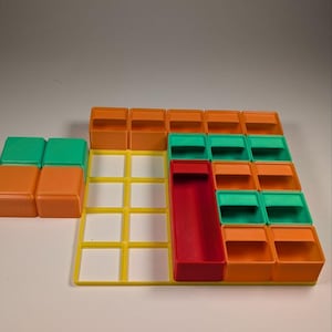 May include: A colorful plastic puzzle game with orange, green, yellow, and red components. The game features a grid-like base with various shaped blocks and containers. The blocks are arranged in a pattern, suggesting a strategy or logic game.