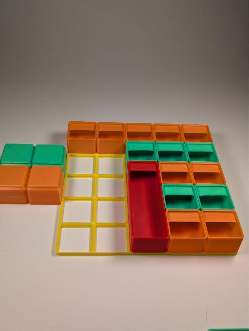 May include: A colorful plastic puzzle game with orange, green, yellow, and red components. The game features a grid-like base with various shaped blocks and containers. The blocks are arranged in a pattern, suggesting a strategy or logic game.