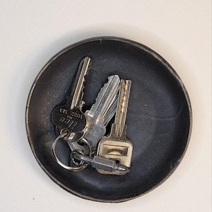 May include: A small, round, matte black ceramic tray containing a metal keyring with three silver-colored keys. One key is clearly marked with the text 1022 SE1 and the brand name ISEO. The tray has a slightly raised rim and a dark, textured finish.