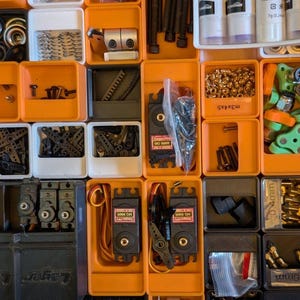 May include: An overhead shot of an organized storage system filled with various small mechanical components. The components include screws, bolts, springs, gears, and electronic parts, all neatly arranged in orange and white compartments.