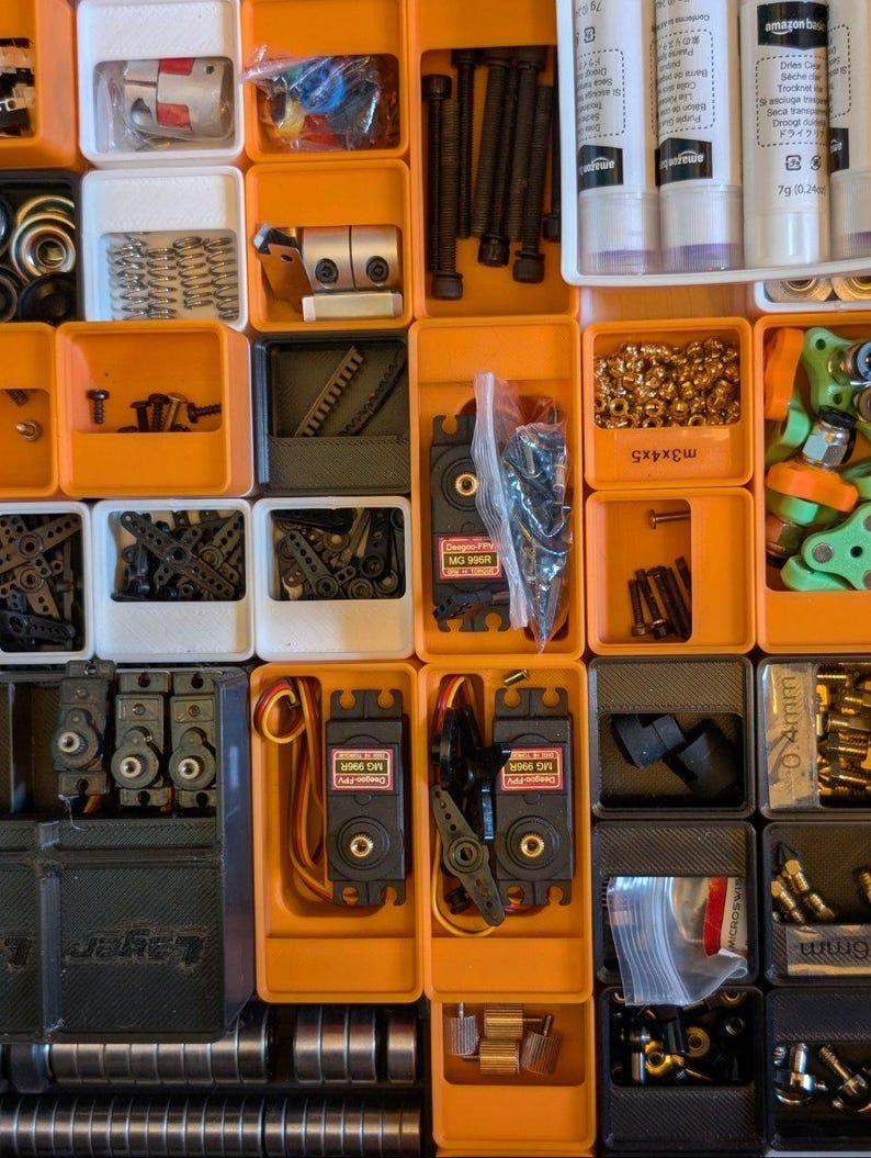 May include: An overhead shot of an organized storage system filled with various small mechanical components. The components include screws, bolts, springs, gears, and electronic parts, all neatly arranged in orange and white compartments.