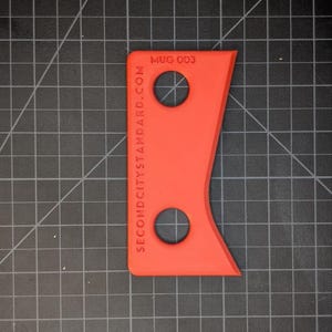 May include: Bright red, uniquely shaped tool with two circular holes. The text "MUG 003" and "SECOND CITY STANDARD.COM" are printed on the tool. The tool is on a black grid surface.