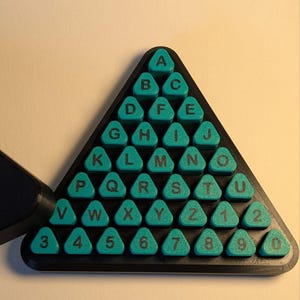 May include: A triangular black base holds a grid of teal-colored triangular tiles. Each tile features a black letter from A to Z or a number from 1 to 0. The tiles are arranged in rows, forming a pyramid shape with the letter A at the top peak.