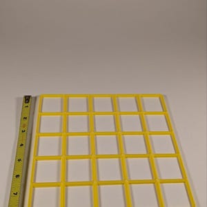 May include: A yellow plastic grid with 25 square openings. A measuring tape is on the left side of the image. The grid is approximately 8 inches wide and 8 inches long. The grid is on a white surface.