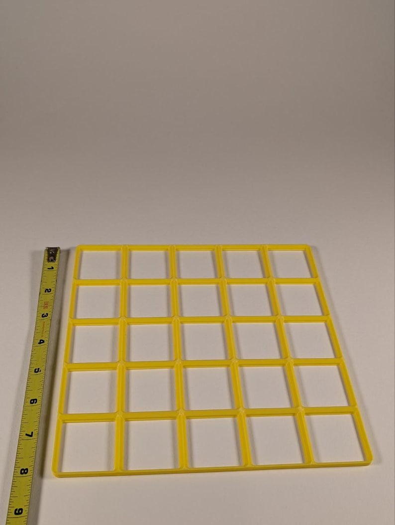 May include: A yellow plastic grid with 25 square openings. A measuring tape is on the left side of the image. The grid is approximately 8 inches wide and 8 inches long. The grid is on a white surface.