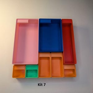May include: A collection of colorful plastic storage containers in various sizes and shapes. The set includes pink, blue, and red rectangular boxes, along with smaller orange and green square containers. The text "Kit 7" is visible.