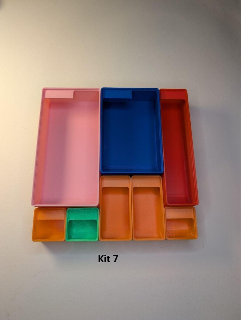 May include: A collection of colorful plastic storage containers in various sizes and shapes. The set includes pink, blue, and red rectangular boxes, along with smaller orange and green square containers. The text "Kit 7" is visible.