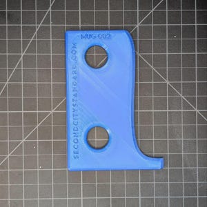 May include: A blue, 3D-printed object with two circular holes and a curved shape. The text "MUG 002" is printed at the top, and "SECONDCITYSTANDARD.COM" is printed along the side. The object is on a black grid surface.