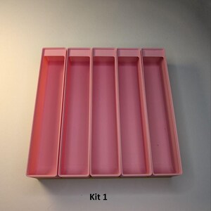 May include: Five pink rectangular storage containers. The containers are arranged side by side, with a slightly raised rim. The text "Kit 1" is printed in black at the bottom of the image. The containers are empty.