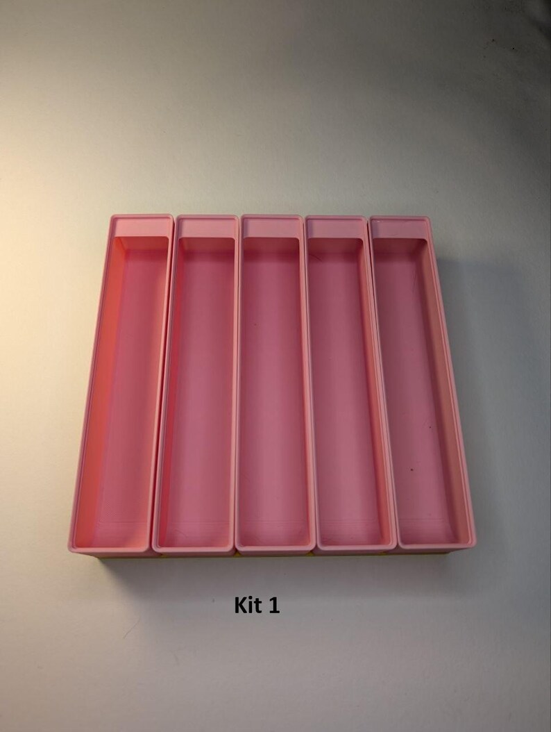 May include: Five pink rectangular storage containers. The containers are arranged side by side, with a slightly raised rim. The text "Kit 1" is printed in black at the bottom of the image. The containers are empty.