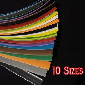 Puede incluir: Un surtido de 12 tiras de colores, dispuestas en forma de abanico sobre un fondo negro. Los colores incluyen tonos de gris, morado, marrón, naranja, amarillo y verde. El texto "12 COLORES" y "10 TAMAÑOS" está en rojo.