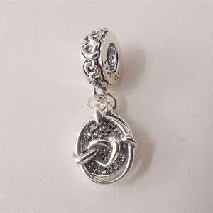 May include: A sterling silver dangle charm featuring a circular pendant with a textured, sparkling center and a raised heart-shaped knot design. The bail is engraved with S 925 ALE and displays a repeating chain-link pattern around its outer edge.