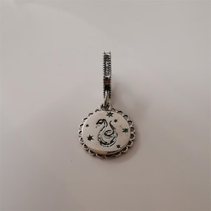 Pode incluir: Berloque prateado com um pendente circular com um desenho de cobra preta e detalhes de estrelas verdes. O berloque tem uma borda decorativa ondulada e uma argola texturizada.