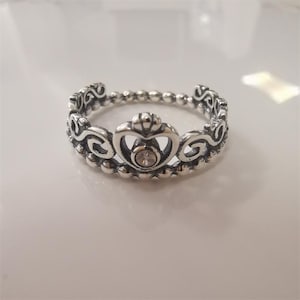 May include: Silver-toned crown ring with a central heart-shaped design and a small, clear gemstone. The ring features a beaded band and ornate scrollwork along the crown's edge, creating a delicate and regal appearance.